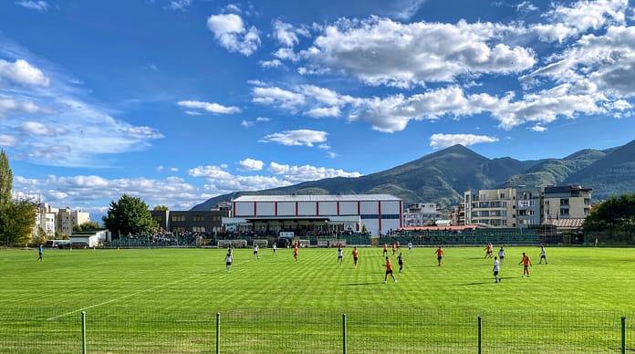 Gotse-Delchev-Bulgaria A local football match taking place on a green pitch in Gotse Delchev, Bulgaria, with mountains and residential buildings in the background.