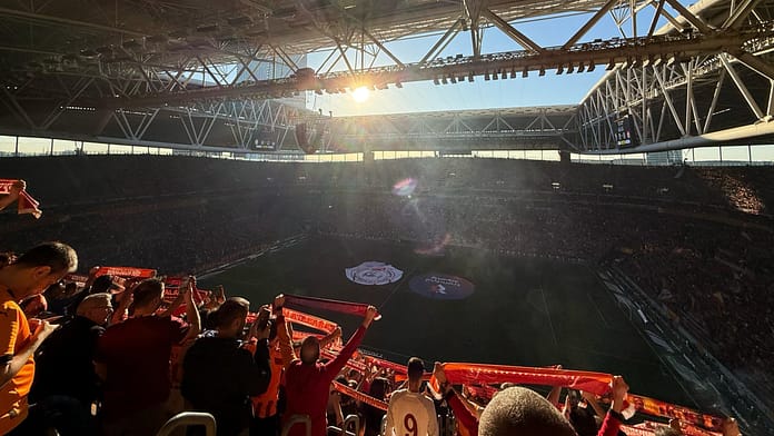 Rams Park - Galatasaray Wide view of Galatasaray fans holding scarves at Rams Park with sunlight streaming over the stadium during a match.