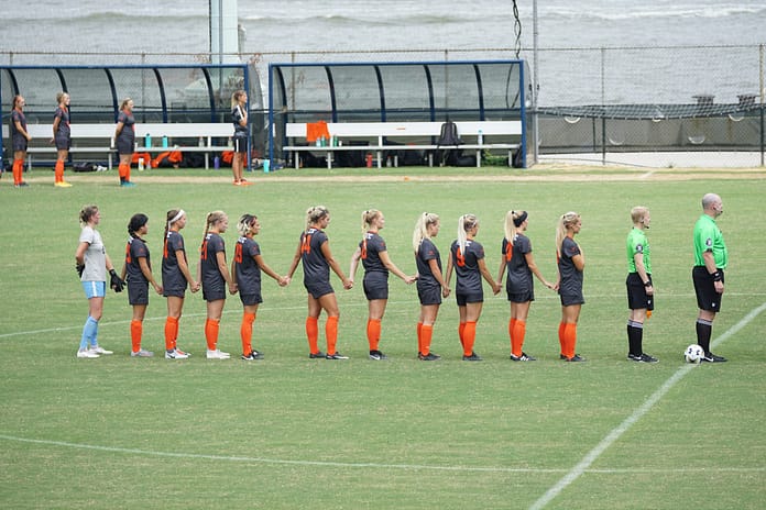 A women’s football team stands hand in hand on the pitch before kickoff, facing the referees during a pre-match ceremony.