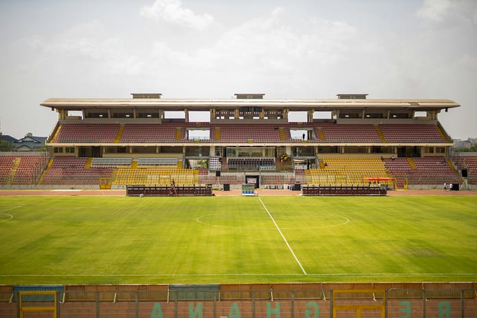 Baba Yara Stadium View of Baba Yara Stadium in Kumasi, Ghana, showing an empty pitch and stands with red, yellow, and white seating under a clear sky.