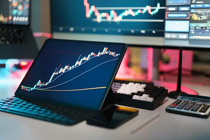 Tablet and monitor displaying financial charts with rising trend lines, alongside a keyboard and calculator on a desk in a trading workspace.