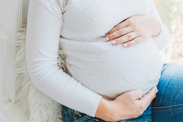 Maternity Pregnant woman in a white sweater and jeans gently holding her baby bump while sitting by a window.