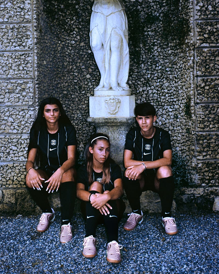 Three F.C. Como Women youth players wearing the 2025/26 Home kit sit in front of a stone statue during The Next Wave campaign shoot.